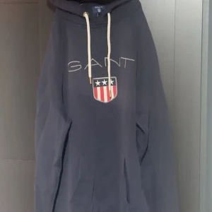 Mörkblå hoodie från GANT - Säljer en snygg och bekväm mörkblå hoodie från GANT. Den har en stor logga på framsidan med ett coolt emblem i rött och vitt. Perfekt för kyliga dagar och har en klassisk känguruficka framtill. Huvan har vita dragsnören för justering. Passar perfekt till vardags eller en avslappnad stil.