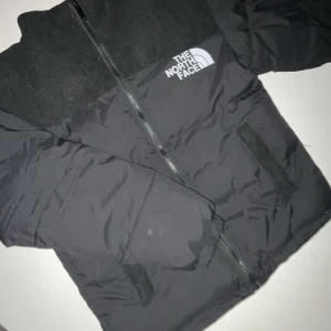 Svart dunjacka från The North Face - Säljer en svart dunjacka från The North Face i mycket bra skick. Jackan har en dragkedja framtill och är perfekt för kalla vinterdagar. Den är långärmad och har en klassisk design med logotypen på bröstet. Supervarm och skön!