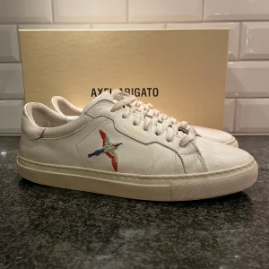 Vita sneakers från Axel Arigato - Säljer ett par stilrena vita sneakers från Axel Arigato i skinn. De har en snygg broderad fågel i blått och rött på sidan och klassisk snörning. Perfekta för både vardag och fest! Skorna är i bra skick med lite slitage på sulan. Kommer med originalkartong.