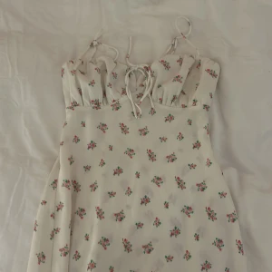 Blommig klänning från Gina Tricot - Charmig blommig klänning från Gina Tricot i en ljus färgton. Klänningen har smala axelband och ett sött knytband framtill. Perfekt för sommarens varma dagar.