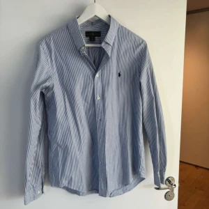 Blå och vit randig skjorta från Ralph Lauren - Snygg blå och vit randig skjorta från Ralph Lauren med klassisk krage och knappar framtill. Skjortan har långa ärmar och en broderad logotyp på bröstet. Perfekt för en stilren look.