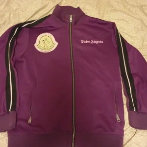 Lila  från Palm Angels×moncler - Säljer en lila jacka från Palm Angels med dragkedja och Moncler-logga på bröstet. Jackan har långa ärmar med svarta och vita ränder längs sidorna. Texten 'MIND CONTROL' finns på baksidan. Perfekt för en unik stil!