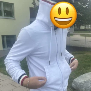 Vit Moncler kofta - Säljer en stilren vit hoodie från Moncler med dragkedja och snörning i huvan. Tröjan har ribbade muddar med röd och blå randdetalj. Perfekt för en casual look. 🏷️