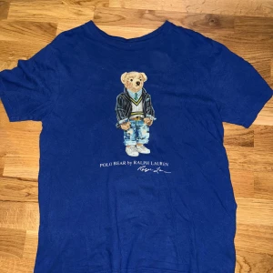 Blå t-shirt med Polo Bear från Ralph Lauren - Säljer en blå t-shirt från Ralph Lauren med det ikoniska Polo Bear-motivet på framsidan. T-shirten har korta ärmar och en rund halsringning. Perfekt för en avslappnad stil.