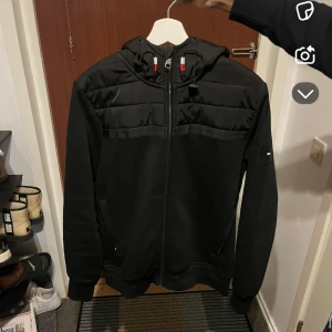 Svart jacka med huva - Säljer en svart jacka med huva och dragkedja. Jackan har långa ärmar och en quiltad design på axlarna. Perfekt för kyligare dagar. Jackan är från Tommy Hilfiger.
