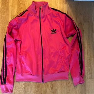 Rosa Adidas kofta - Säljer en snygg rosa träningsjacka/kofta från Adidas med svarta ränder.  Adidas-loggan finns både fram och bak.