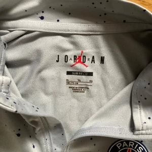 Grå långärmad tröja från Jordan x PSG - Säljer en grå långärmad tröja från Jordan i samarbete med PSG. Tröjan har ett stänkmönster och en halv dragkedja framtill. Den har också PSG-loggan och texten 'Accor Live Limitless' på bröstet. Perfekt för sportiga tillfällen!