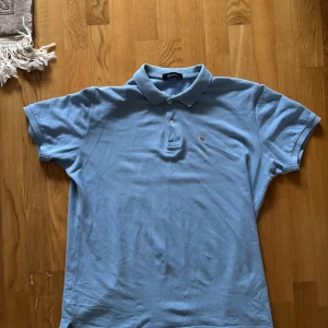 Ljusblå pikétröja från GANT - Säljer en stilren ljusblå pikétröja från GANT med klassisk krage och knappar. Tröjan har en broderad logotyp på bröstet och är perfekt för en avslappnad look.