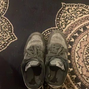 Grå Nike Air Force 1 - Säljer ett par grå Nike Air sneakers med snörning och perforerade detaljer på ovansidan. Skorna har en klassisk Nike-logga i silver på sidan och en robust sula för extra komfort.