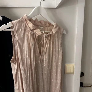 Ljusrosa ärmlös topp från H&M - Säljer en söt ljusrosa ärmlös topp från H&M med diskret mönster och knytband vid halsen. Perfekt för varma dagar och ger en romantisk touch till din stil. 💕