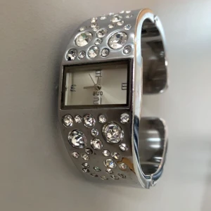 Glittrig silverklocka från One - Snygg och glittrig klocka från One i silver med gnistrande stenar på armbandet. Den har en rektangulär urtavla med stilrena visare och markeringar. Perfekt för att lägga till lite bling till din outfit! ✨