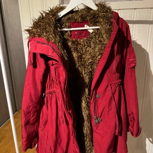 Röd parkajacka från Odd Molly - Säljer en snygg röd parkajacka från Odd Molly med fuskpälsfoder. Jackan har dragkedja och knappar framtill samt justerbar midja för en perfekt passform. Perfekt för kyliga dagar!