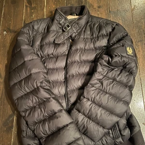 Svart pufferjacka från Belstaff - Säljer en snygg svart pufferjacka från Belstaff. Jackan har en dragkedja framtill och en hög krage med spänne. Perfekt för kyliga, våriga och höstiga dagar med sin quiltade design. Märkeslogga på ärmen ger en extra touch av stil. Väldigt varsamt använd inga märken och ser ut o va ny skick. Ny pris 4300