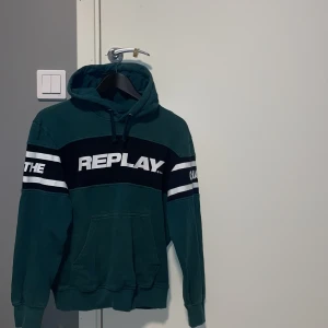 Grön hoodie från Replay - Snygg grön hoodie från Replay med svart och vit text på bröstet. Den har en klassisk känguruficka och justerbar huva med snören. Perfekt för en avslappnad stil. OBS FÄRGEN PÅ TRÖJAN SER INTE UT SÅDÄR! Mer bilder kan skickas vid köp!