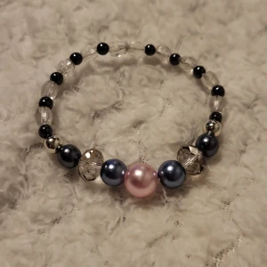 Färgglatt pärlarmband - Charmigt armband med en mix av pärlor i olika färger. Det har en stor rosa pärla i mitten, omgiven av blå och svarta pärlor samt genomskinliga och silverfärgade detaljer. Perfekt för att ge en färgklick till din outfit.