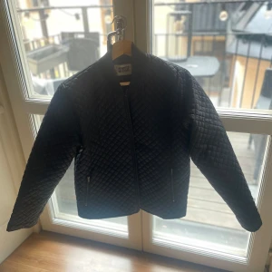 Svart quiltad jacka från Weekday - Snygg svart quiltad jacka från Weekday i storlek medium. Jackan har en dragkedja framtill och en stilren design med långa ärmar. Perfekt för en cool och avslappnad look.