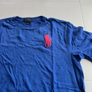 Blå t-shirt från Polo Ralph Lauren - Säljer en blå t-shirt från Polo Ralph Lauren med en stor röd broderad logga på bröstet och en röd siffra på ärmen. Perfekt för en avslappnad stil. T-shirten är kortärmad och tillverkad i mjukt material.