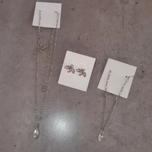Smyckesset  i silver - Elegant set med ett halsband och matchande armband och örhängen  i silver. Halsbandet har en fin kedja med en droppformad hängande detalj såsom armbandet. Örhängena är dekorerade med små, glittrande stenar som ger en lyxig känsla.