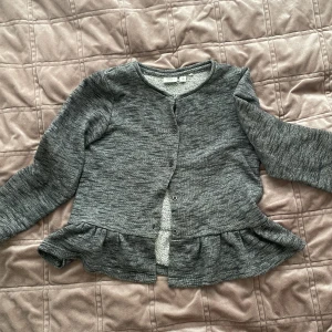 Grå kofta från Name It - Säljer en söt grå kofta från Name It med volangdetalj nedtill. Koftan har långa ärmar och knappar framtill. 