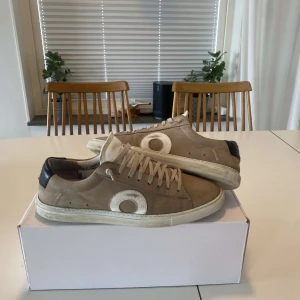 Beige sneakers från Oliver Cabell - Snygga beige sneakers från Oliver Cabell med vita detaljer och snörning. Skorna har en stilren design med en rundad tå och en kontrasterande svart häl. Perfekta för en avslappnad stil. Hör av er vid funderingar. Pris kan diskuteras. Box medföljer.
