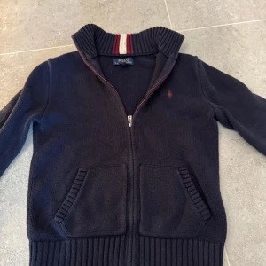Mörkblå stickad kofta från Ralph Lauren - Säljer en snygg mörkblå stickad tröja från Ralph Lauren med dragkedja och ribbad krage. Tröjan har två praktiska fickor framtill och en logga på bröstet. Perfekt för den kommande våren! Tveka inte på att fråga! /Vikram 
