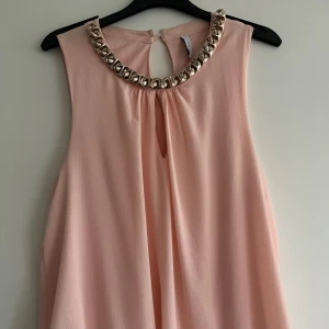 Ljusrosa ärmlös topp från Amisu - Säljer en elegant ljusrosa ärmlös topp från Amisu med dekorativa silverdetaljer runt halsen. Toppen har en liten öppning bak med knappstängning. Perfekt för en stilren look.