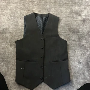 Svart väst kostymväst - Snygg svart väst med knappar framtill och justerbart spänne bak. Perfekt för en stilren look. Något stor över bysten på mig därav säljer jag