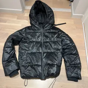 Vi har fått möjligheten att få in en svart pufferjacka för tjejer. Den kommer från Stradivarius med huva och dragkedja. Jackan har en glansig yta och justerbara snören i nederkant för en bättre passform. Perfekt för kyliga dagar.jackan är i nyskick