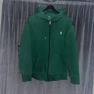 Grön hoodie från Polo Ralph Lauren - Säljer en snygg grön hoodie från Polo Ralph Lauren med dragkedja och den klassiska loggan broderad i vitt på bröstet. Perfekt för en avslappnad stil med justerbar huva och fickor framtill. Passar bra till både jeans och joggers.
