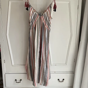 Randig sommarklänning från Lindex - Lång randig och klockad klänning från Lindex i olika färger som beige, röd, vit, grå och svart. Klänningen har smala justerbara axelband med dekorativa tofsar och en luftig passform. Rundad och vid nertill. 