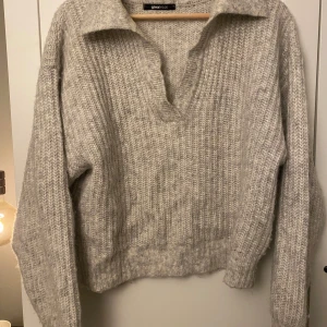 Grå stickad tröja från Gina Tricot - Mysig grå stickad tröja från Gina Tricot med en avslappnad passform och en stilren krage. Tröjan har långa ärmar och en mjuk textur som gör den bekväm att bära.