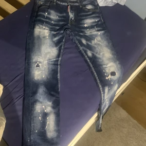 Blå jeans från Dsquared2 - Säljer ett par coola blå jeans från Dsquared2 med slitna detaljer och färgstänk. De har en normal passform och är perfekta för en avslappnad stil. Jeansens unika design gör dem till ett statement-plagg i garderoben.