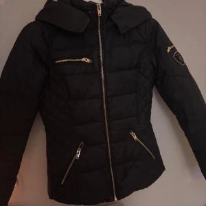 Svart jacka från Rockandblue - Snygg svart quiltad jacka från Rockandblue med dragkedjor och huva. Jackan har en stilren design med flera praktiska fickor och ett emblem på ärmen. Säljes utan päls då ja tappat bort den samt smink vid kragen men de går bort vid tvätt. Nypris ca 2500kr