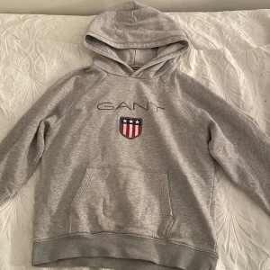 Grå hoodie från GANT - Säljer en grå hoodie från GANT med ett snyggt emblem på bröstet. Tröjan har en stor ficka framtill och en mjuk insida som håller dig varm. Perfekt för kyliga dagar!