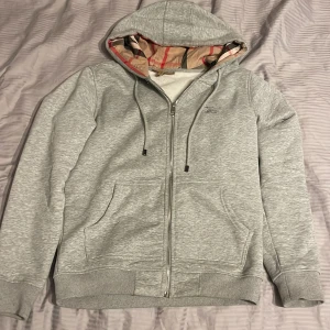 Grå hoodie  Burberry - Säljer en stilren grå hoodie från Burberry med dragkedja och justerbar huva. Insidan av huvan har det klassiska Burberry-mönstret i beige, svart och rött. Tröjan har broderad logga på bröstet och praktiska fickor framtill.