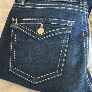 Mörkblå jeans med kontrastsömmar - Snygga mörkblå jeans med vita kontrastsömmar och en klassisk femficksdesign. Jeansen har en knappdetalj på bakfickan och är perfekta för en avslappnad stil.