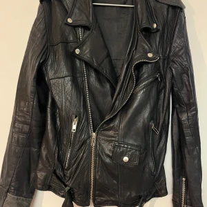 Svart skinnjacka från Deadwood - Snygg svart skinnjacka från Deadwood med coola dragkedjedetaljer och knappar. Jackan har en klassisk bikerstil med flera fickor och en asymmetrisk dragkedja framtill. Perfekt för att ge din outfit en edgy touch.