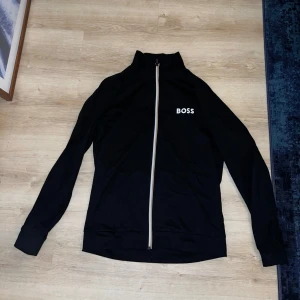 Svart tröja från BOSS - Säljer en stilren svart tröja från BOSS med dragkedja framtill. Jackan har långärmade ärmar och en hög krage. Perfekt för en casual look.