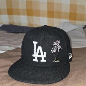 Svart keps från New Era - Snygg svart keps från New Era med LA-logga framtill och broderade detaljer som palmer och ett hjärta. På sidan finns ett märke för Dodger Stadiums 50-årsjubileum. Perfekt för att ge din outfit en cool touch.