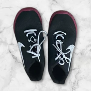 Svarta Nike sneakers med röd sula - Säljer ett par svarta Nike sneakers med vit swoosh och röd sula. Skorna har vita snören och en sportig design, perfekt för en aktiv livsstil.