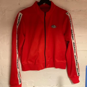 Röd jacka från Dickies - Snygg klarröd jacka från Dickies med dragkedja och logga på bröstet samt på ärmarna. Superbra layer-plagg, säljer pga för kort i ärmarna! Ingen storlek angiven men skulle säga xs. Har en missfärgning på ena ärmen (syns på bilden) men annars bra skick! Köpt på humana!