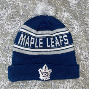 Blå mössa med Toronto Maple Leafs-logga - Snygg blå mössa med vit text 'MAPLE LEAFS' och en grå tofs på toppen. Mössan har en broderad Toronto Maple Leafs-logga framtill och NHL-logga baktill. Perfekt för vintern och för fans av laget!