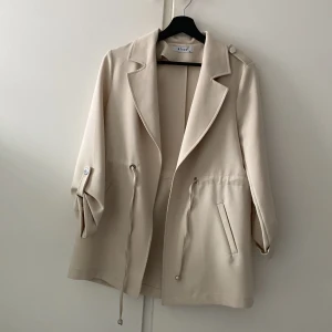 Beige trenchcoat från Klapa - Snygg beige trenchcoat från Klapa med knytband i midjan och knappar på axlarna. Jackan har en klassisk krage och långa ärmar med knappdetaljer. Perfekt för vår och höst!