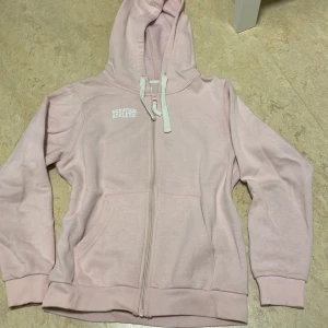 Ljusrosa hoodie från Nonation Athletic - Säljer en ljusrosa hoodie från Nonation Athletic med dragkedja och justerbar huva. Perfekt för en avslappnad stil. Den har en tryckt logga på bröstet och ribbade muddar vid ärmslut och nederkant.Använd max 3-5 gånger. Köpt på stadium. För liten för mig