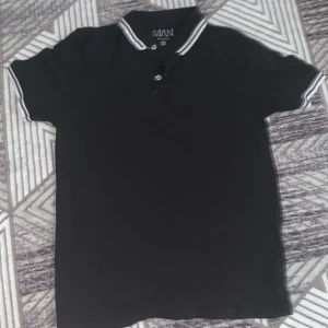 Svart pikétröja från boohooMAN - Säljer en stilren svart pikétröja från boohooMAN. Tröjan har korta ärmar och en klassisk krage med vita ränder. Perfekt för en avslappnad men ändå snygg look.