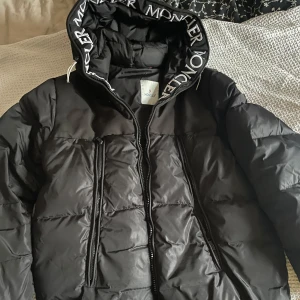 Svart dunjacka från Moncler - Snygg svart dunjacka från Moncler med logotyp på huvan. Jackan har dragkedja framtill och är perfekt för kyligare väder. Den är tillverkad i polyester och har en fyllning av dun för extra värme. Den är givetvis äkta! Nypris:26.000