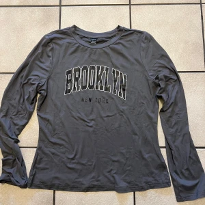 Grå långärmad tröja med Brooklyn-tryck - Snygg grå långärmad tröja med texten 'Brooklyn New York' tryckt på framsidan. Perfekt för en avslappnad stil och bekväm att bära. Passar bra till jeans eller joggers.
