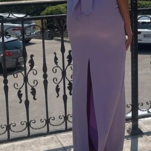 Ljuslila långklänning med pärldetaljer - Elegant ljuslila långklänning med smala axelband och v-ringning dekorerad med pärlor. Klänningen har en slits baktill och en figurnära passform som ger en stilren siluett. Perfekt för speciella tillfällen. 