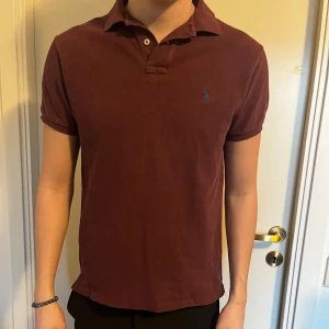 Vinröd pikétröja från Ralph Lauren - Säljer en stilren vinröd pikétröja från Ralph Lauren med klassisk krage och knappdetalj. Tröjan har korta ärmar och en liten broderad logotyp på bröstet. Perfekt för en avslappnad men ändå snygg look.