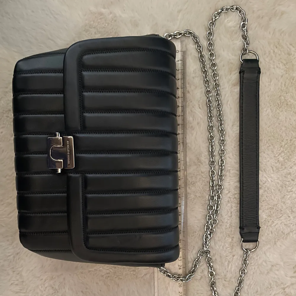Snygg svart axelväska i quiltat skinn från By Malene Birger. ❤️ Väskan har en elegant kedjerem och ett stilrent spänne framtill. Insidan är fodrad i en kontrasterande röd färg, vilket ger en lyxig känsla. Perfekt för att lyfta din outfit! 🤩Mycket sparsamt använd. Spänningen har ett par små skrapor, knappt synliga.. Laukut & Käsilaukut.
